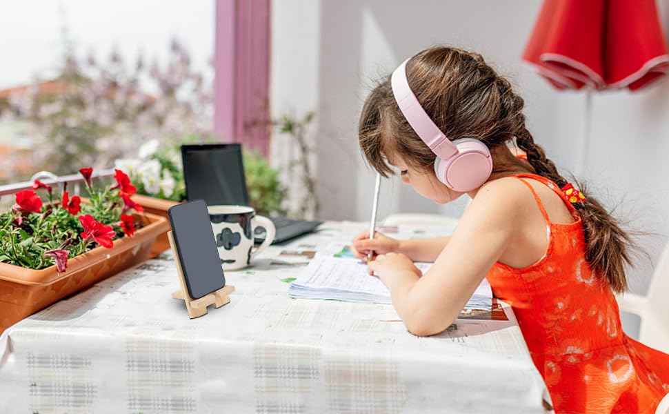 Child in red dress writing or drawing at outdoor table with laptop and flowerpot nearby, demonstrating portable work or study setup.