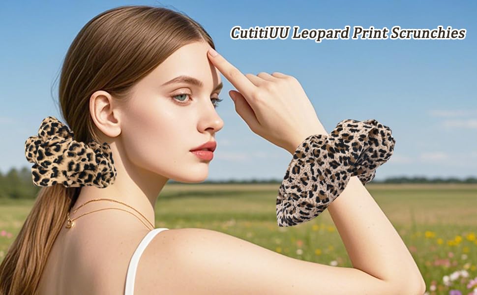 CutiiUU leopard print scrunchies worn on wrist and in hair. Large, fluffy hair accessories with tan and black spotted pattern against outdoor field backdrop.