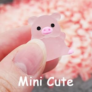 A small, pink, pig-shaped silicone or rubber item held between thumb and forefinger. The background is blurred with pink tones.